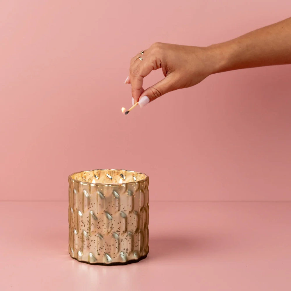 A hand lighting a multi-wick candle in a textured gold glass jar against a solid pink background.