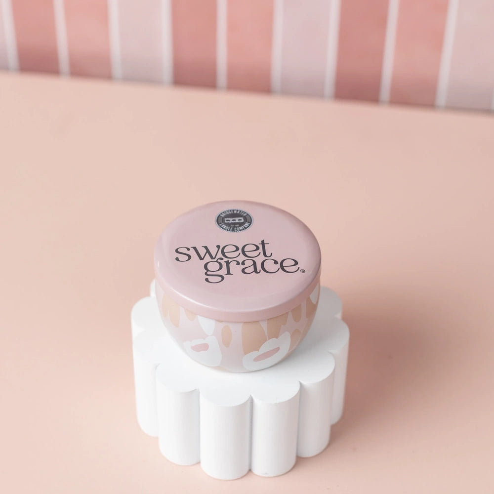 Sweet Grace small candle tin sitting atop a white scalloped pedestal against a pink background.