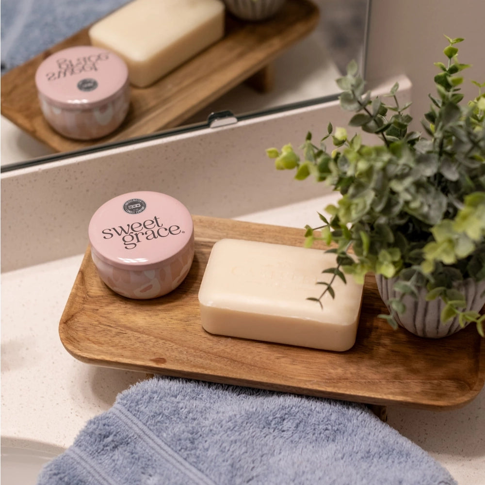 Sweet Grace tin candle, bar soap, and potted plant displayed on a wooden tray in a bathroom setting.