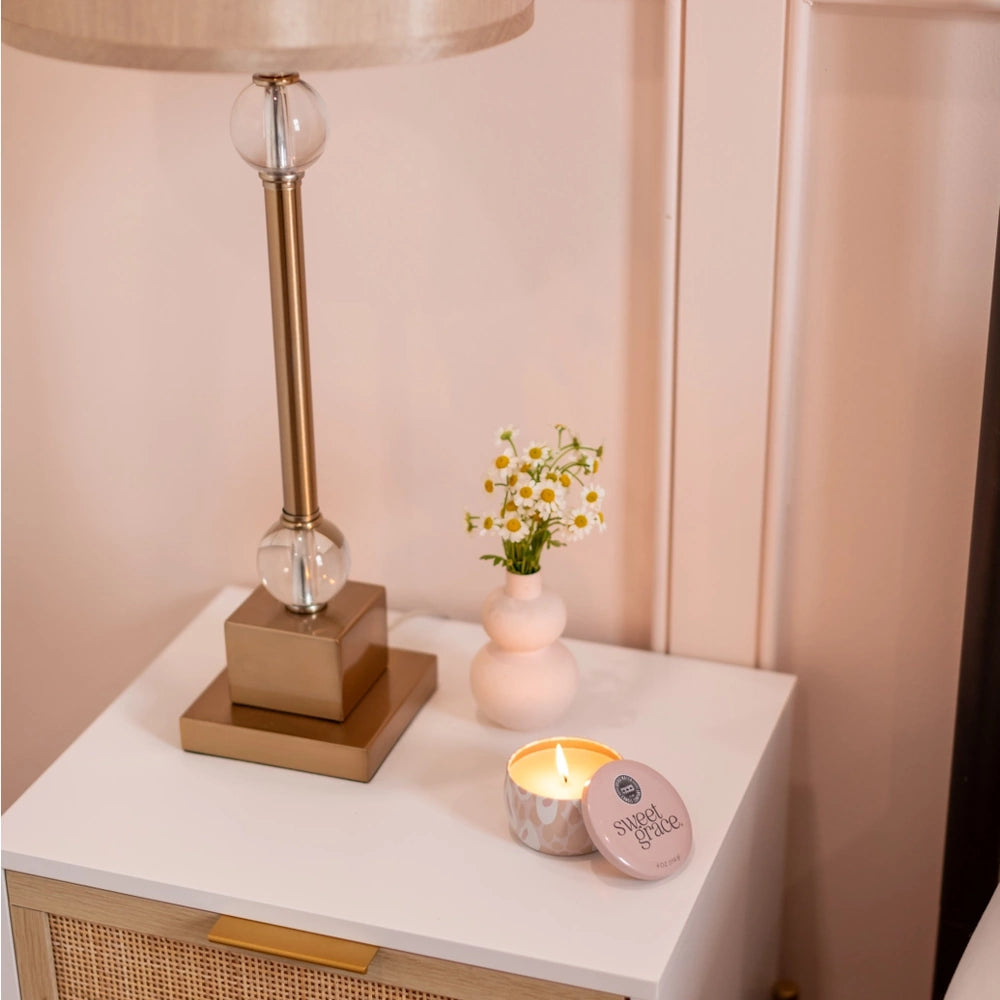 Lit candle in a patterned tin on a white nightstand next to a lamp and small vase of flowers.