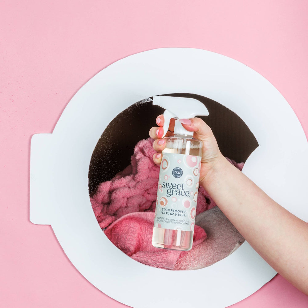 Hand spraying Sweet Grace Stain Remover bottle into a washing machine door with pink laundry inside.