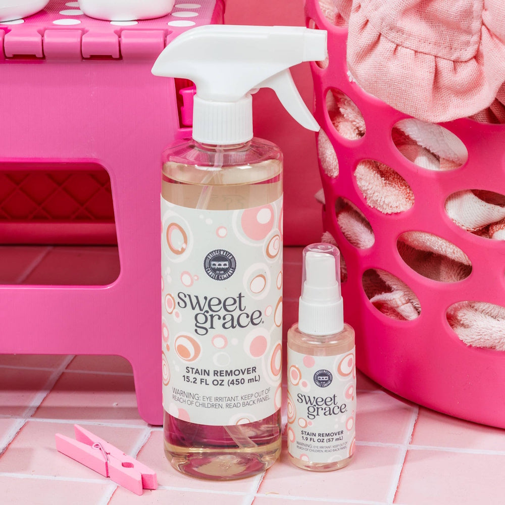 Two Sweet Grace stain remover spray bottles on pink tile, next to a pink laundry basket and clothespin.