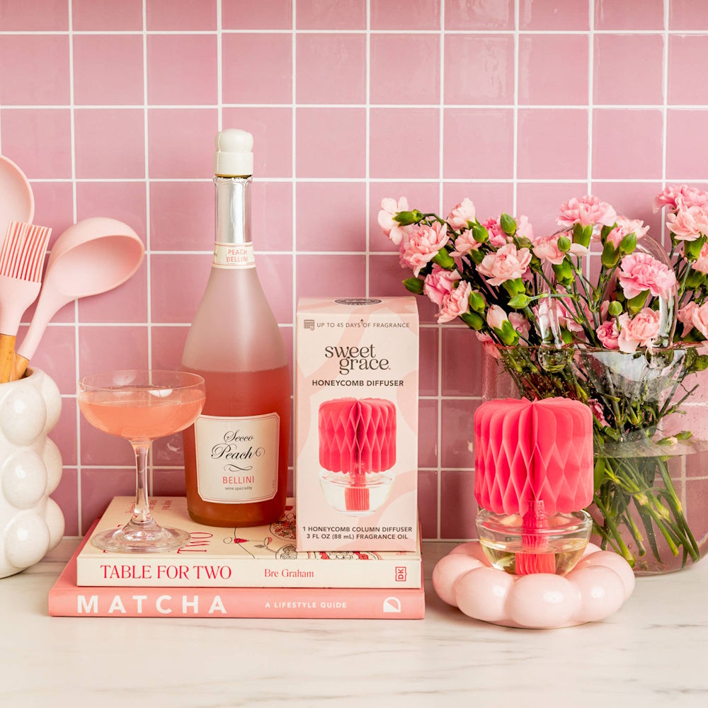 Sweet Grace Honeycomb Diffuser and box next to a Bellini bottle, cocktail glass, and pink flowers.