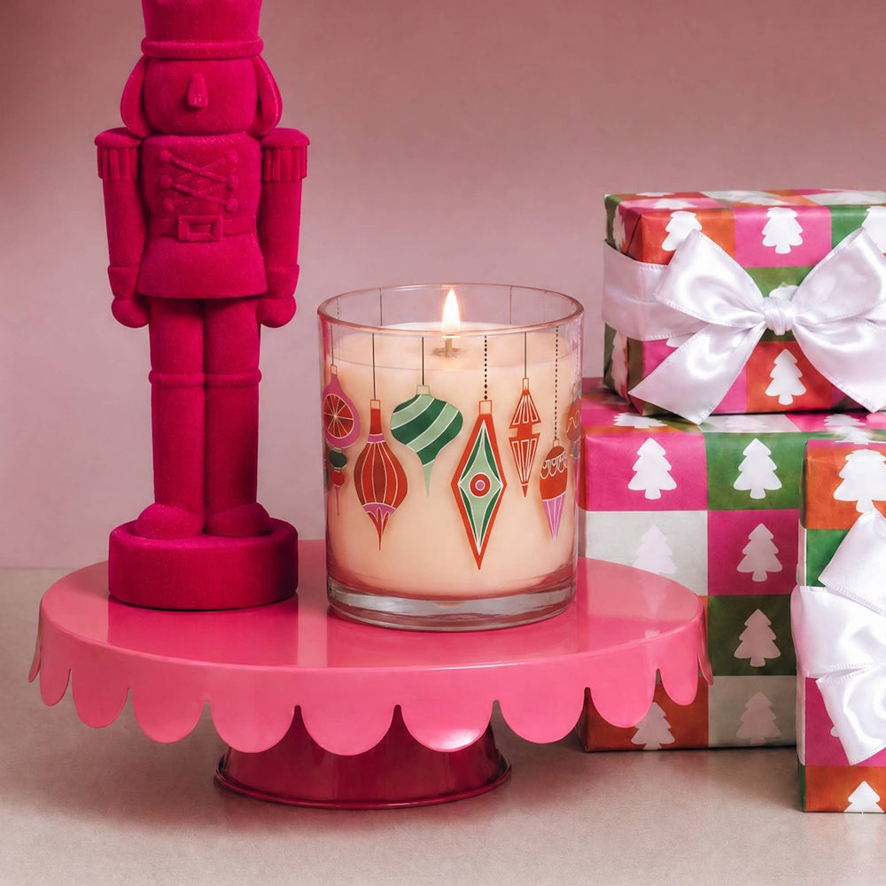 Lit ornament glass candle on a pink stand, next to a pink velvet nutcracker and wrapped gifts.