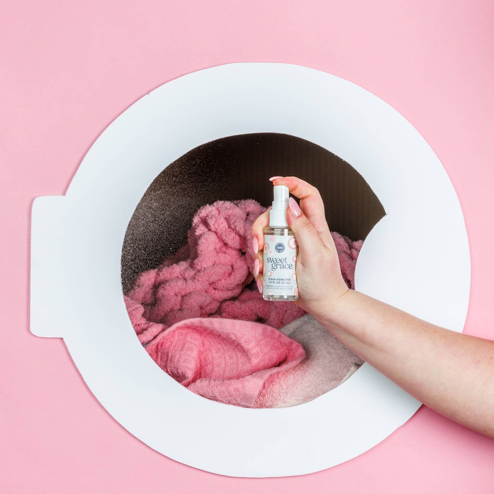 Hand holding a mini Sweet Grace Stain Remover bottle inside a stylized washing machine frame with pink laundry.