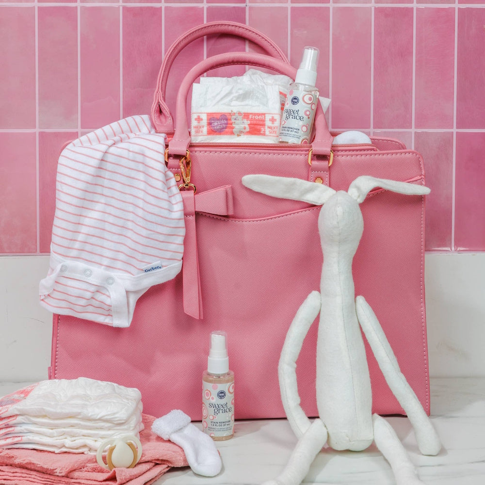 Pink purse filled with baby items, including a Sweet Grace stain remover spray, diapers, and a stuffed bunny.