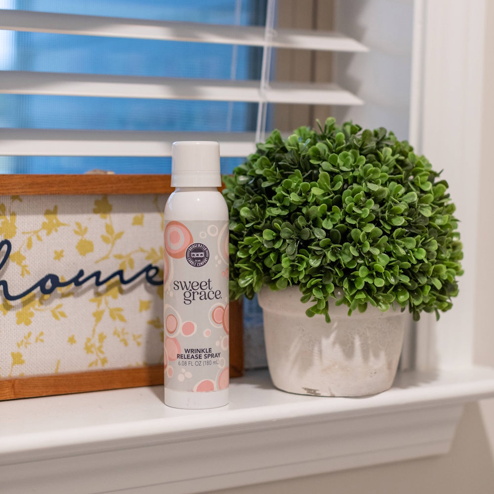 Sweet Grace wrinkle release spray bottle on a windowsill next to a potted green plant.