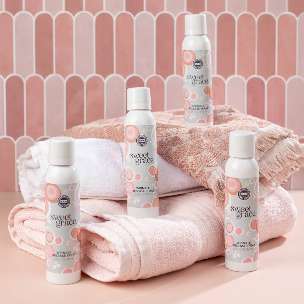 Four bottles of Sweet Grace wrinkle release spray arranged around stacked folded pink towels.