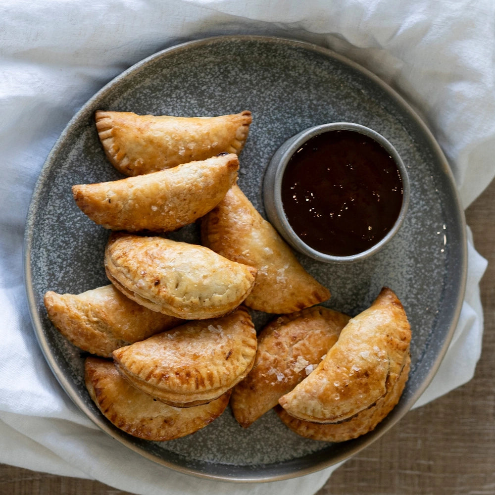 Golden brown baked empanadas served on a grey plate with a side of dark dipping sauce and a white linen background.