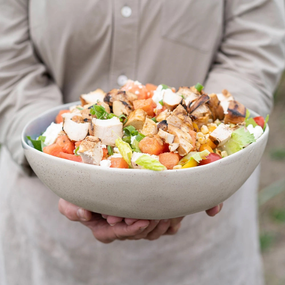 Grilled chicken salad bowl with tomatoes, greens, and cheese held by a person.
