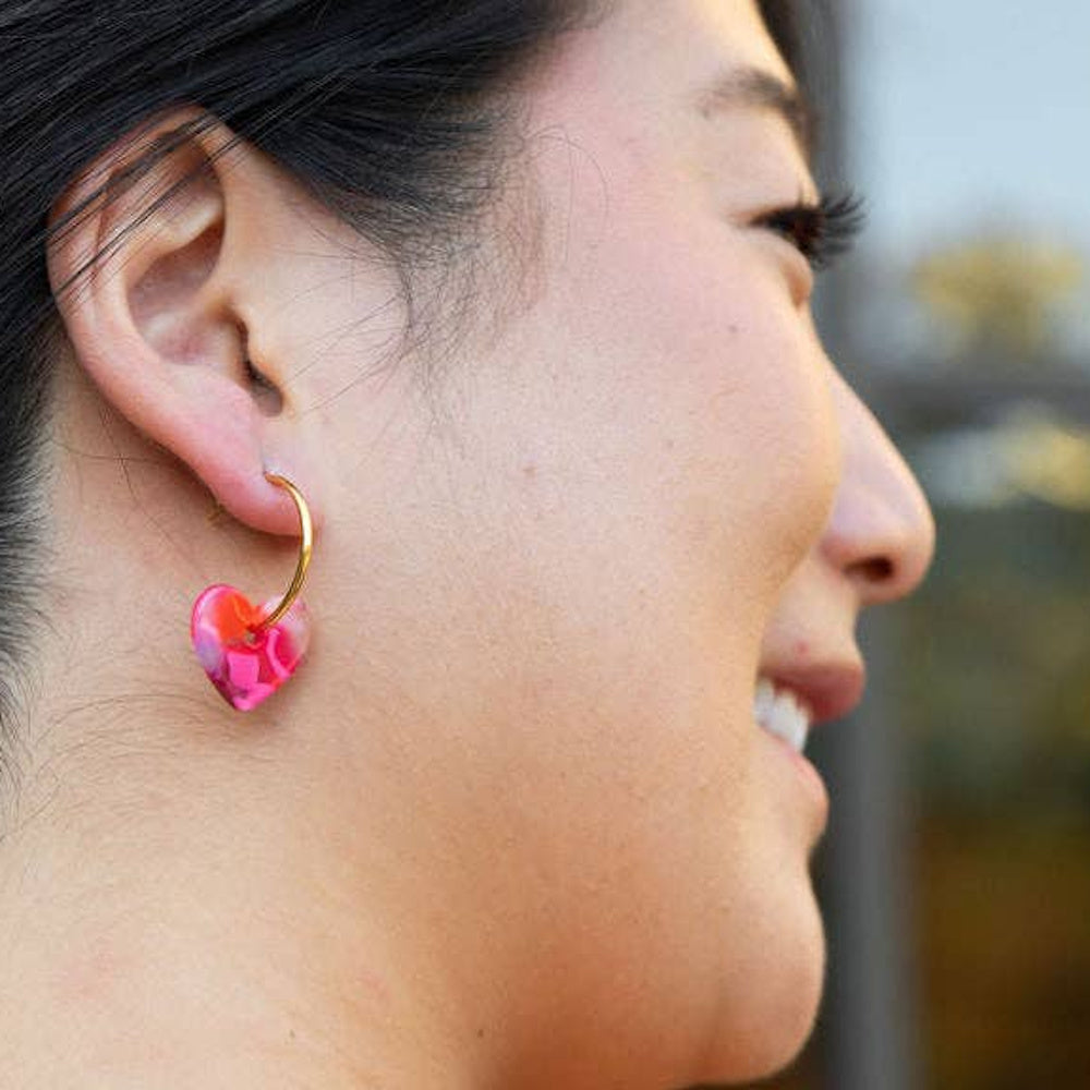 Smiling woman wearing a gold hoop earring with a pink marbled resin heart charm.
