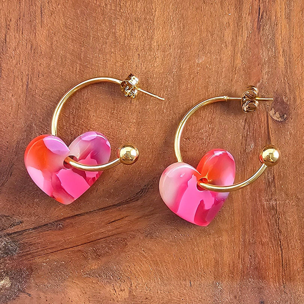 Gold hoop earrings with marbled pink and orange resin heart charms on a wood background.