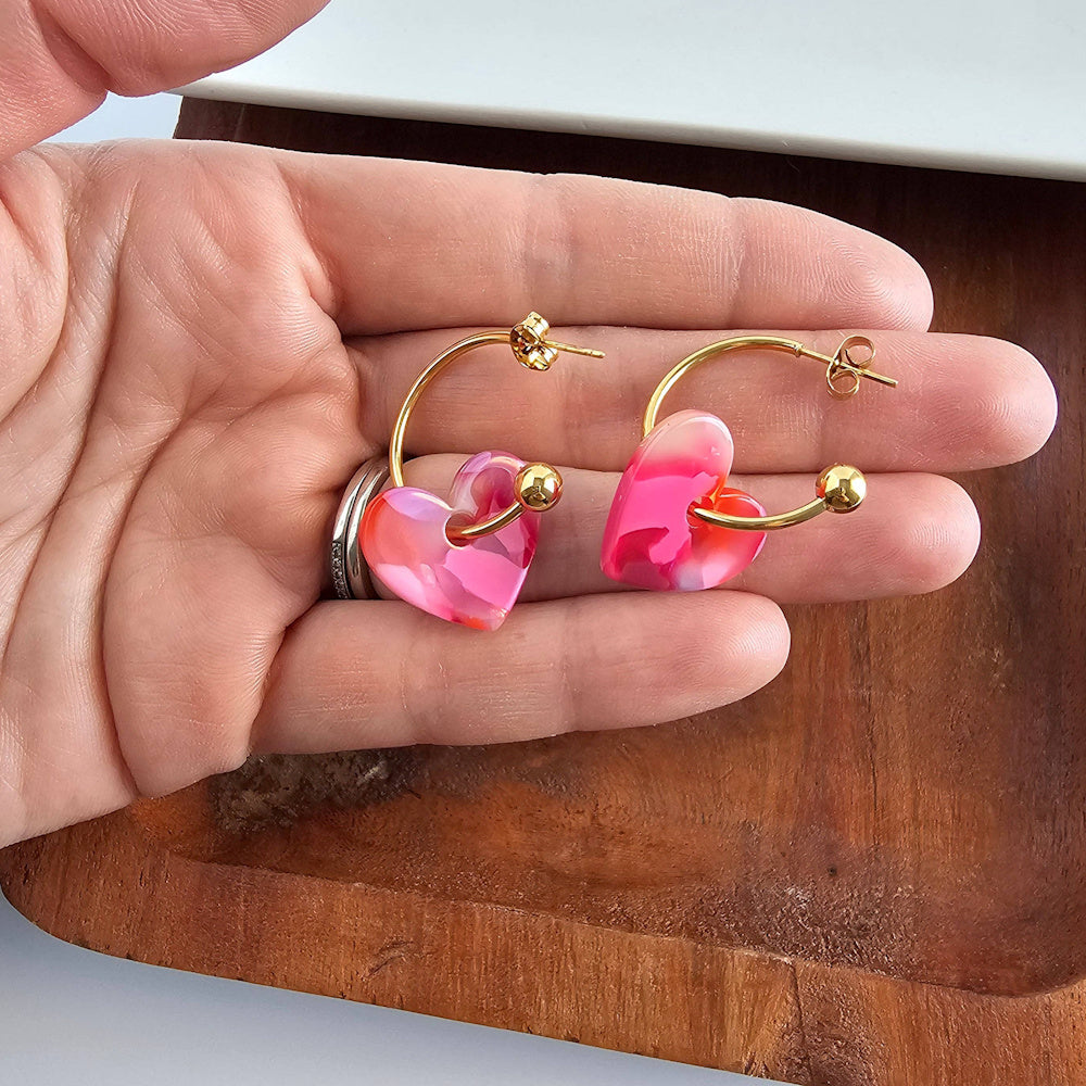 Pink marble heart charms on gold hoop earrings held in a hand over a wooden tray.