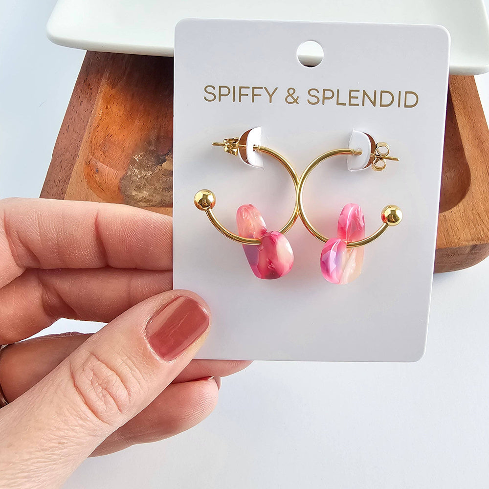 Spiffy & Splendid gold Sweetheart Hoops with pink marble heart charms on a white jewelry card, held by a hand.