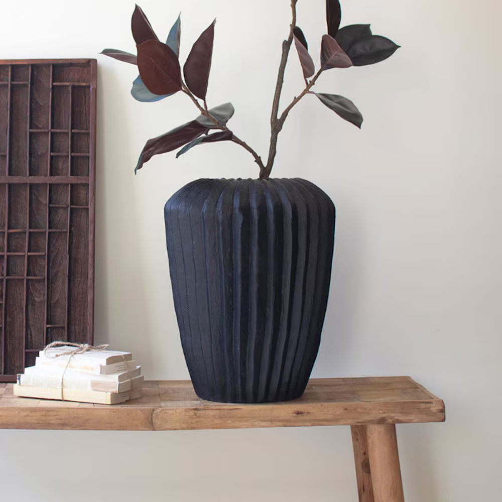 Black fluted stone composite urn vase with a textured surface holding a branch on a rustic wooden bench.