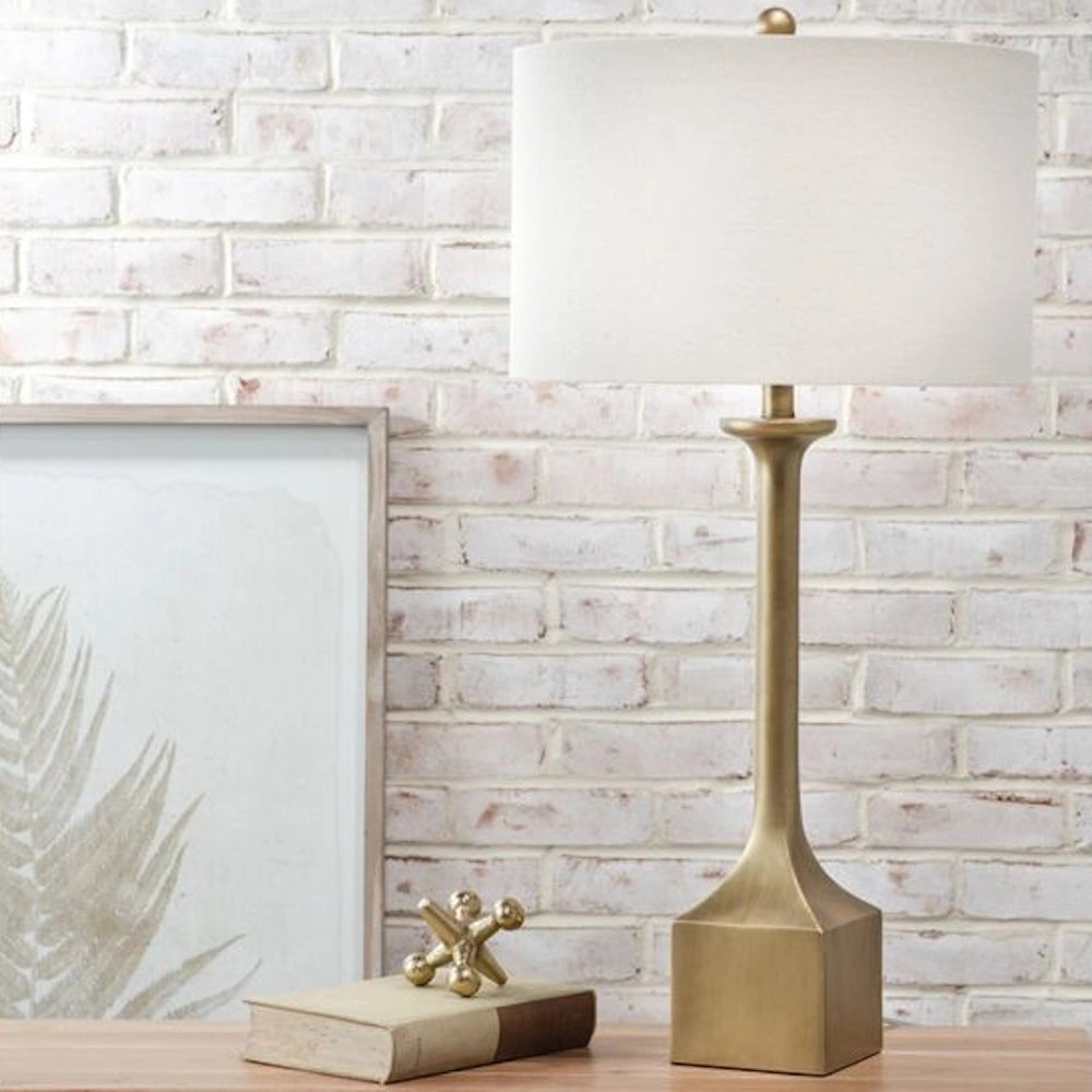 Tall gold resin table lamp with a square base and white drum shade on a wooden surface against a white brick wall.