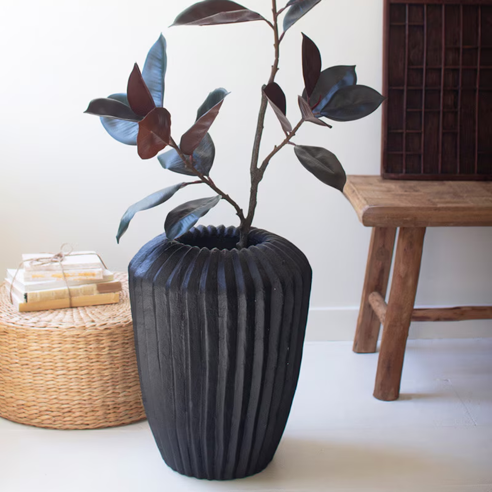 Black fluted stone composite urn planter holding a rubber plant, styled with a woven ottoman and wooden bench.