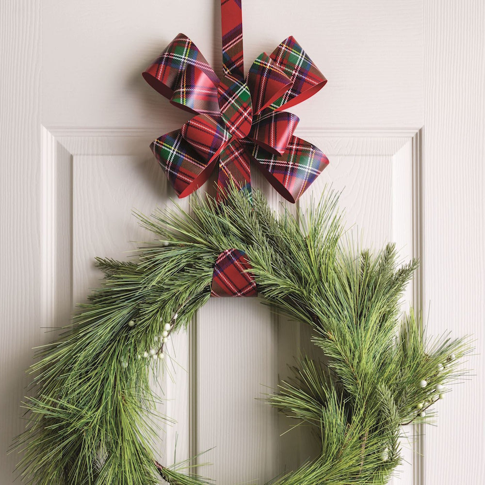 Close-up of a red tartan plaid metal bow hanger holding a pine wreath on a white door.