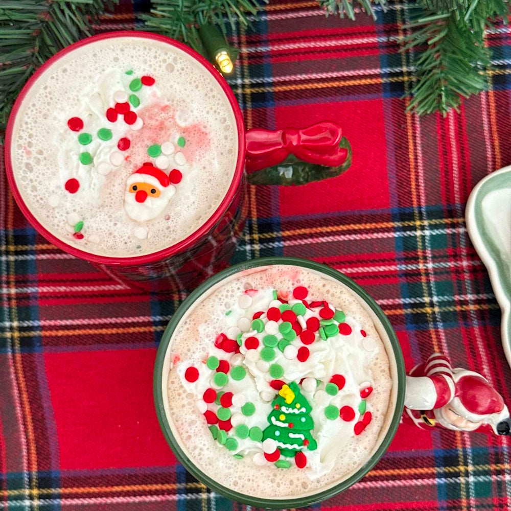 Top-down view of holiday mugs filled with hot cocoa, whipped cream, sprinkles, and festive toppers.