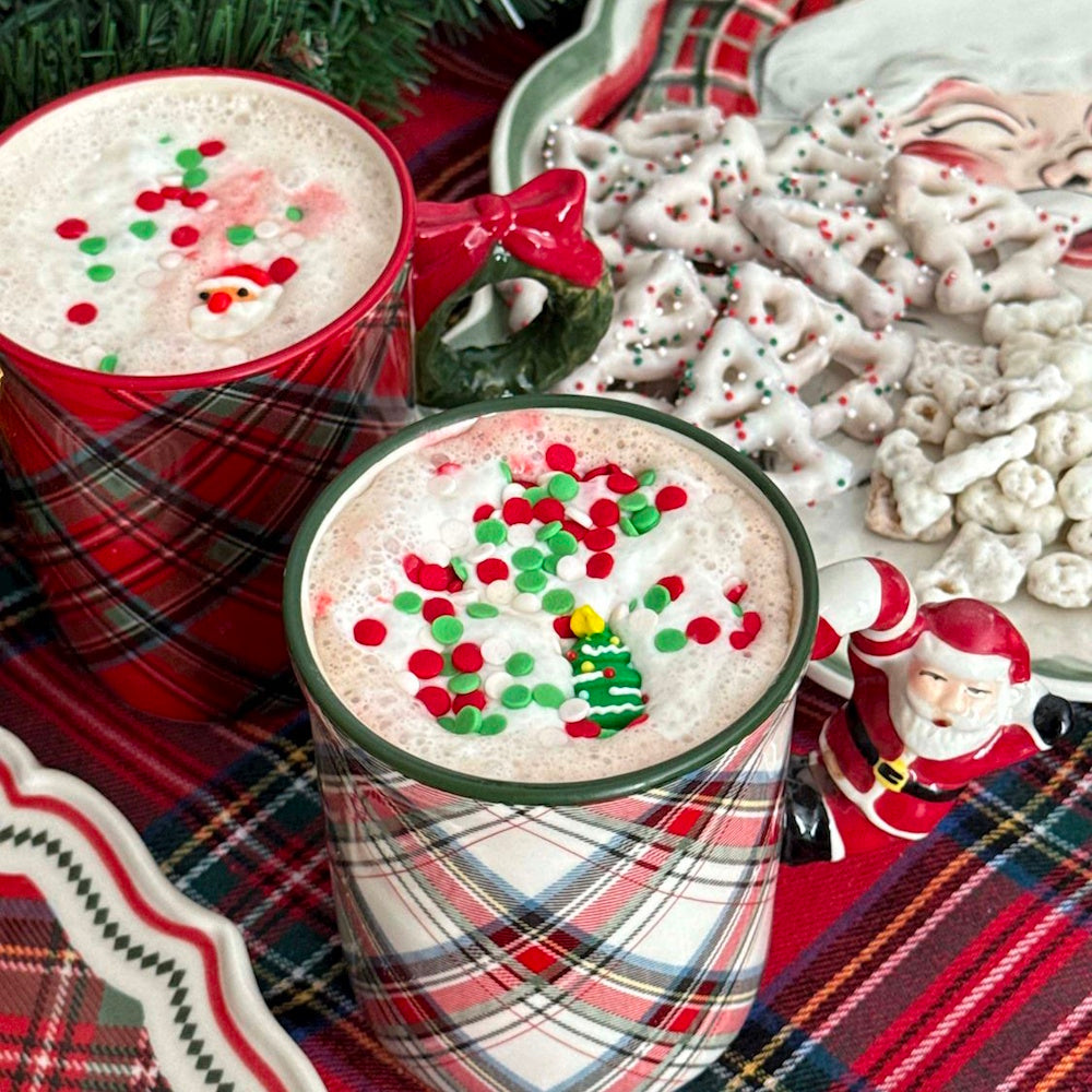 Two tartan plaid mugs filled with hot cocoa and festive sprinkles, featuring Santa and wreath handles.