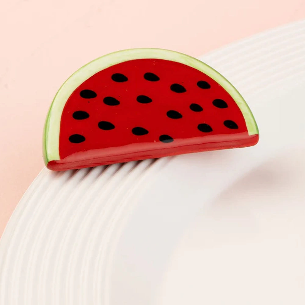 A glossy, semi-circular watermelon slice accessory with a green rind and black seeds on a white plate.