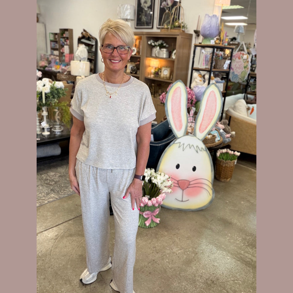 Woman in a taupe short-sleeve knit shirt and matching pants set posing in a home decor boutique with Easter decorations.