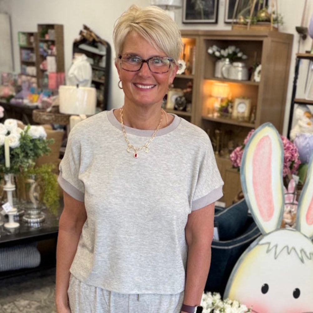 Smiling woman with short blonde hair and glasses wearing a beige short-sleeve knit top in a boutique shop.