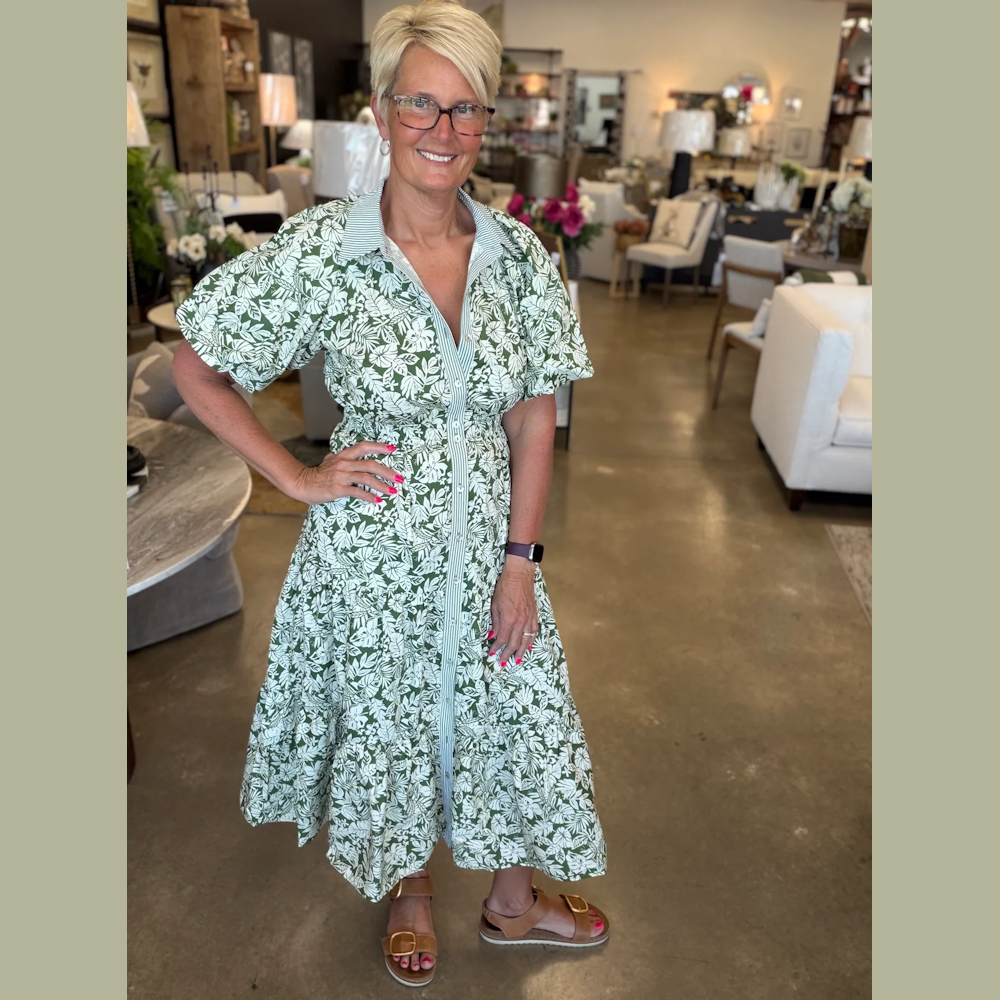 A woman with short blonde hair wears a green leaf-print tiered midi dress and tan sandals in a furniture store.