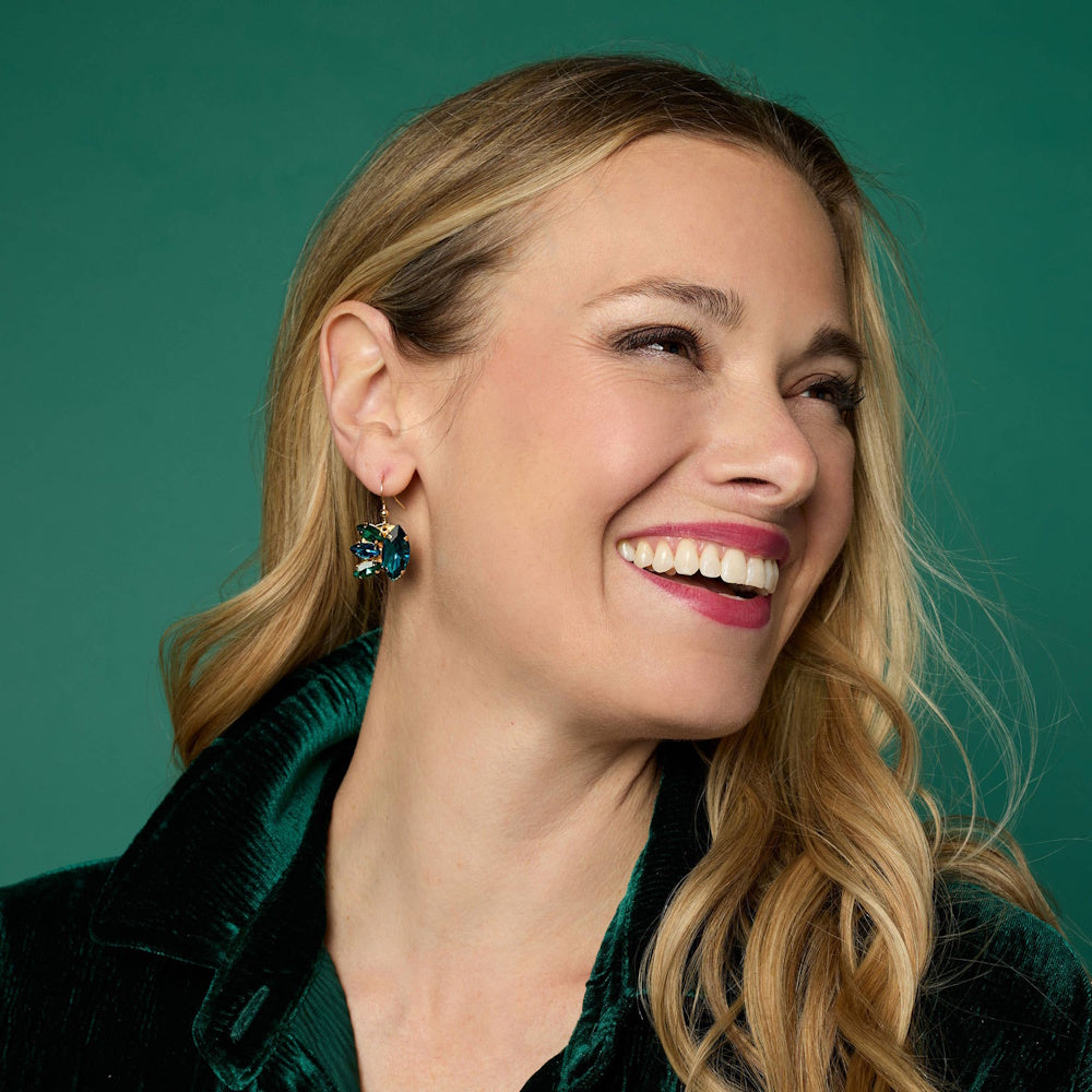 Smiling woman looking away, wearing a deep green velvet jacket and a teal jewel burst dangle earring.