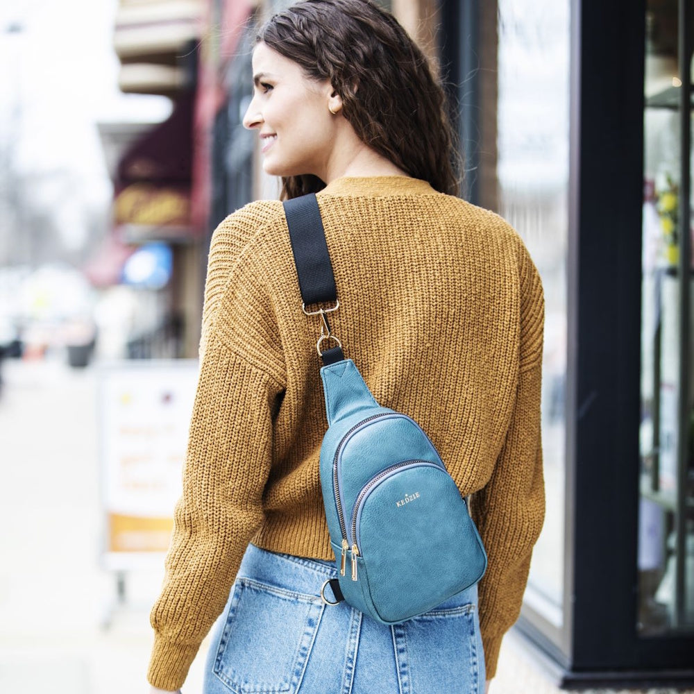 Woman in a mustard yellow sweater wearing a blue sling bag across her back while walking outdoors.