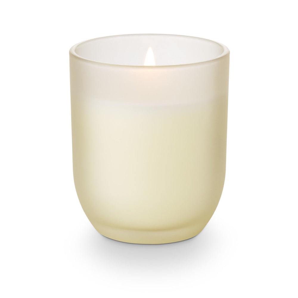 Lit scented candle in a frosted cream glass jar, glowing softly against a white background.