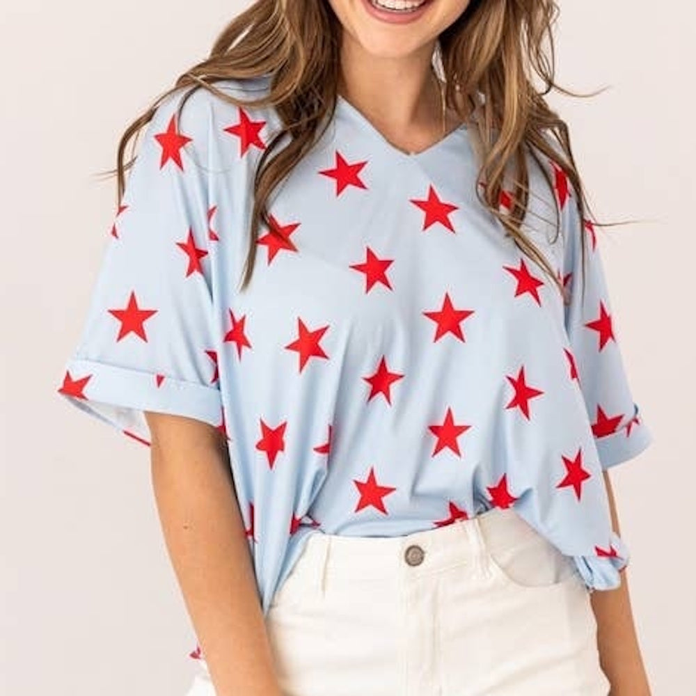A woman wearing a light blue V-neck t-shirt with a red star pattern, tucked into white pants.