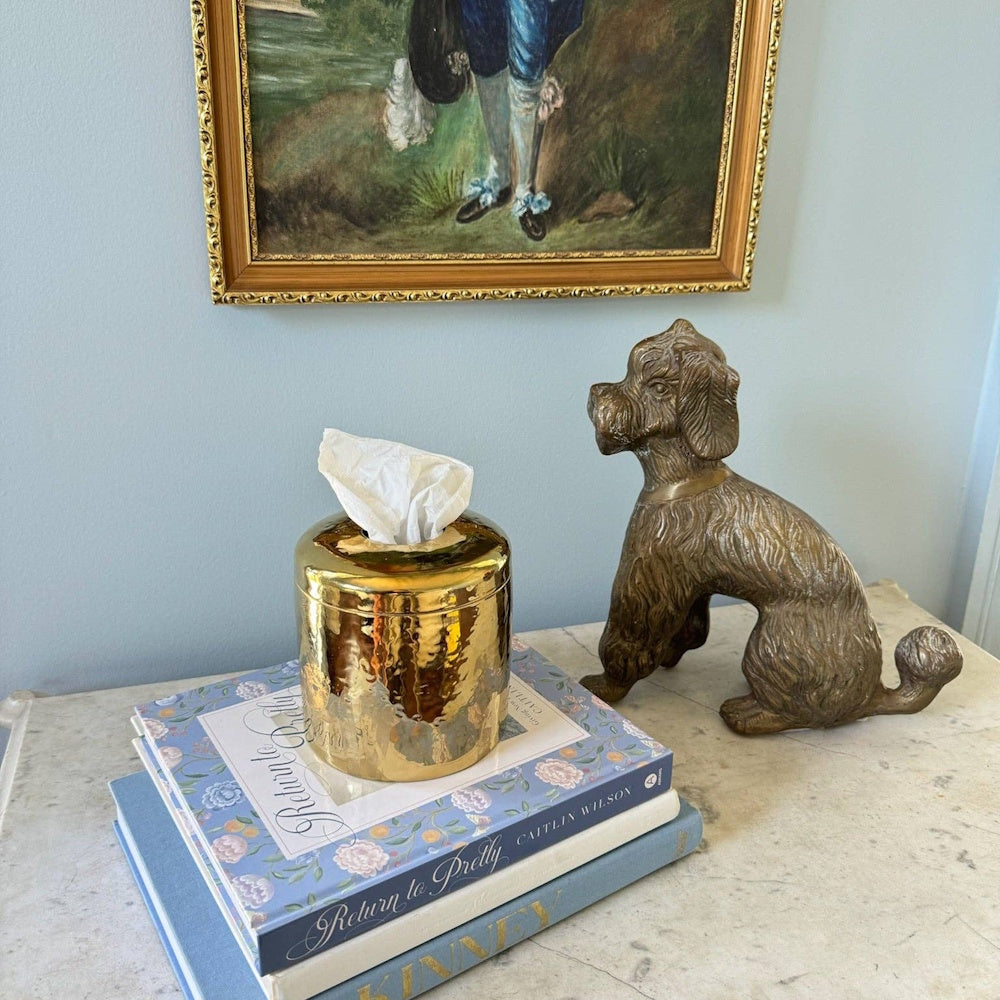 Gold hammered tissue dispenser on stacked books next to a brass dog figurine and framed art.