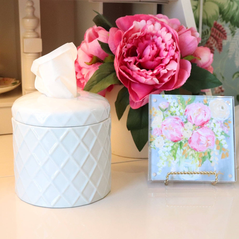 White textured tissue jar next to pink peonies and a small floral painting on a white surface.