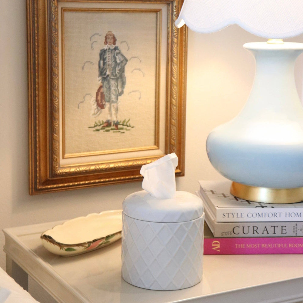A white ceramic tissue holder sits on a bedside table next to books and a blue lamp with an ornate gold frame.