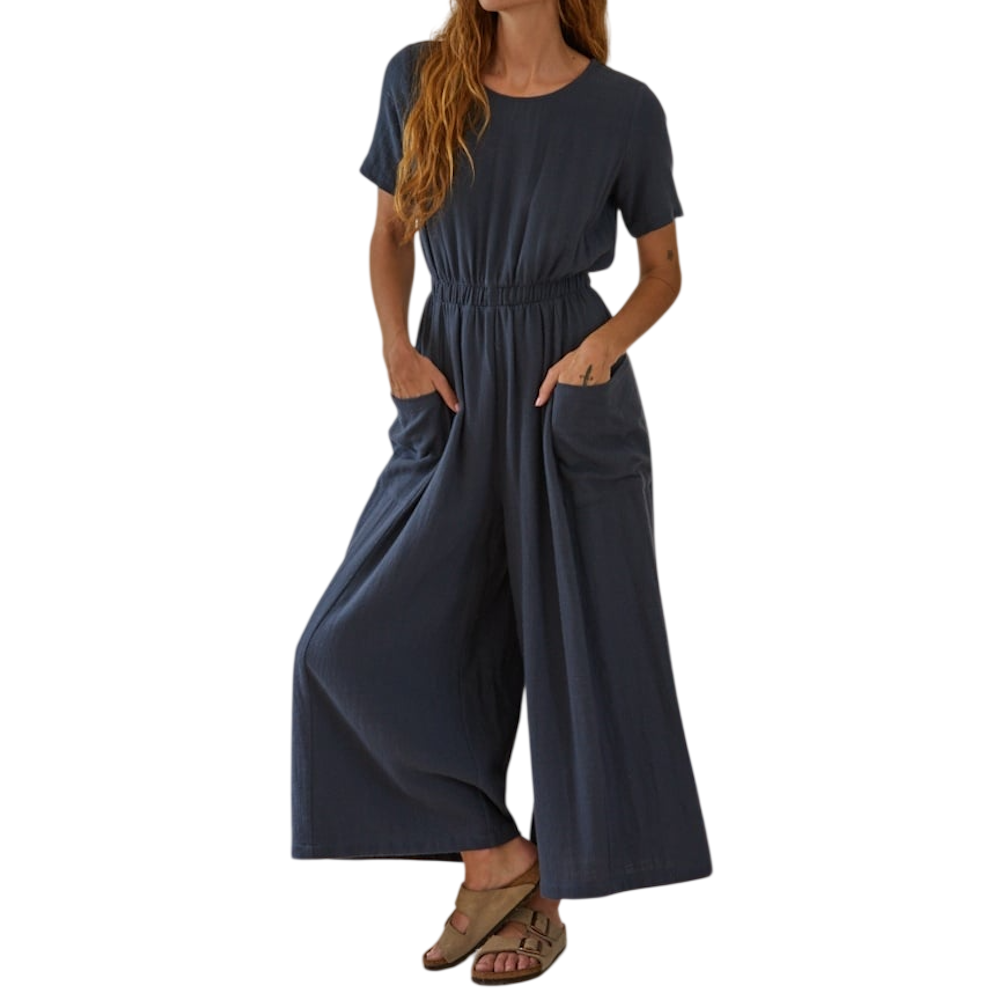 A person wearing a short-sleeved, wide-leg blue jumpsuit with an elastic waist, pockets, and sandals.