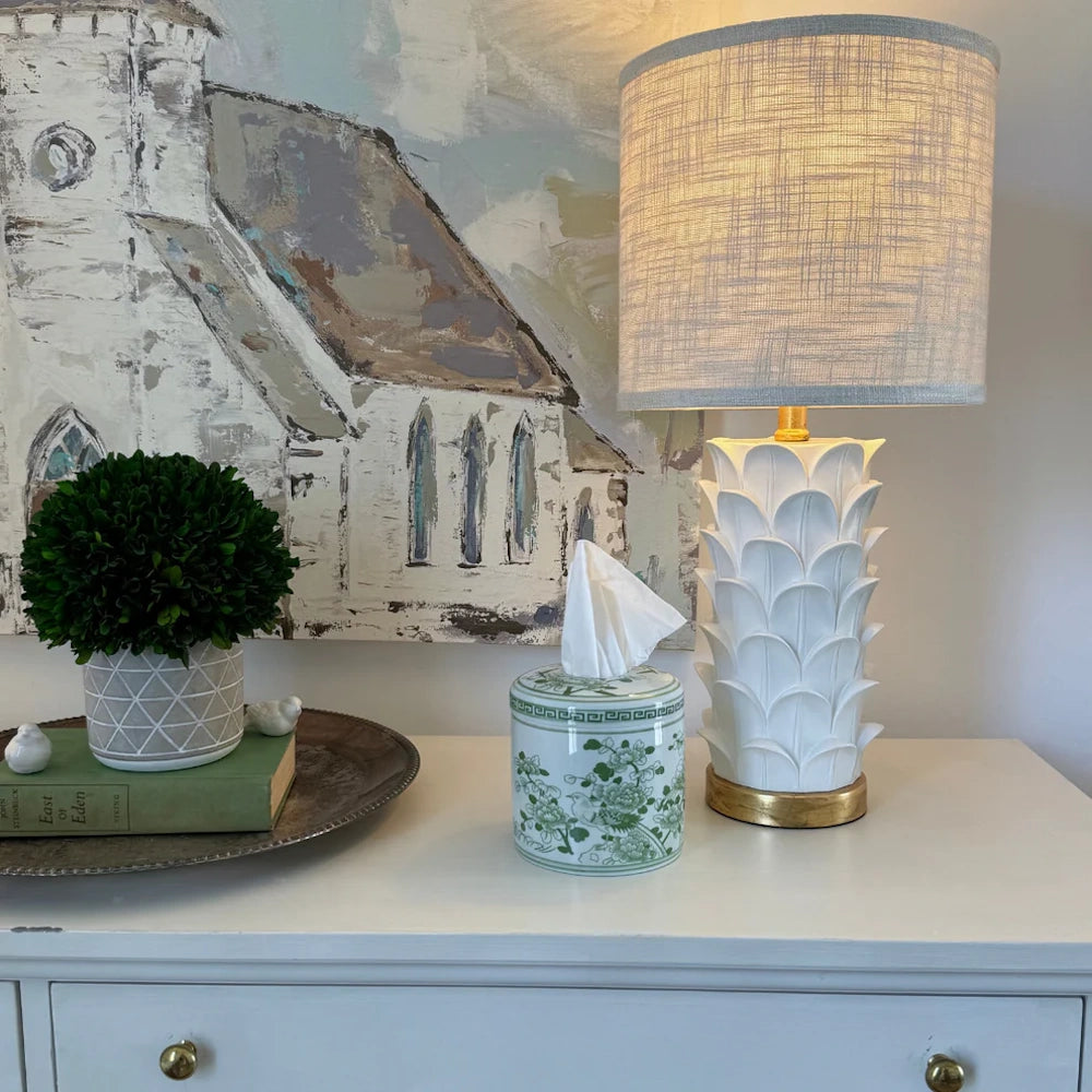 Green and white floral ceramic tissue box cover on a white dresser with an artichoke lamp and landscape painting.