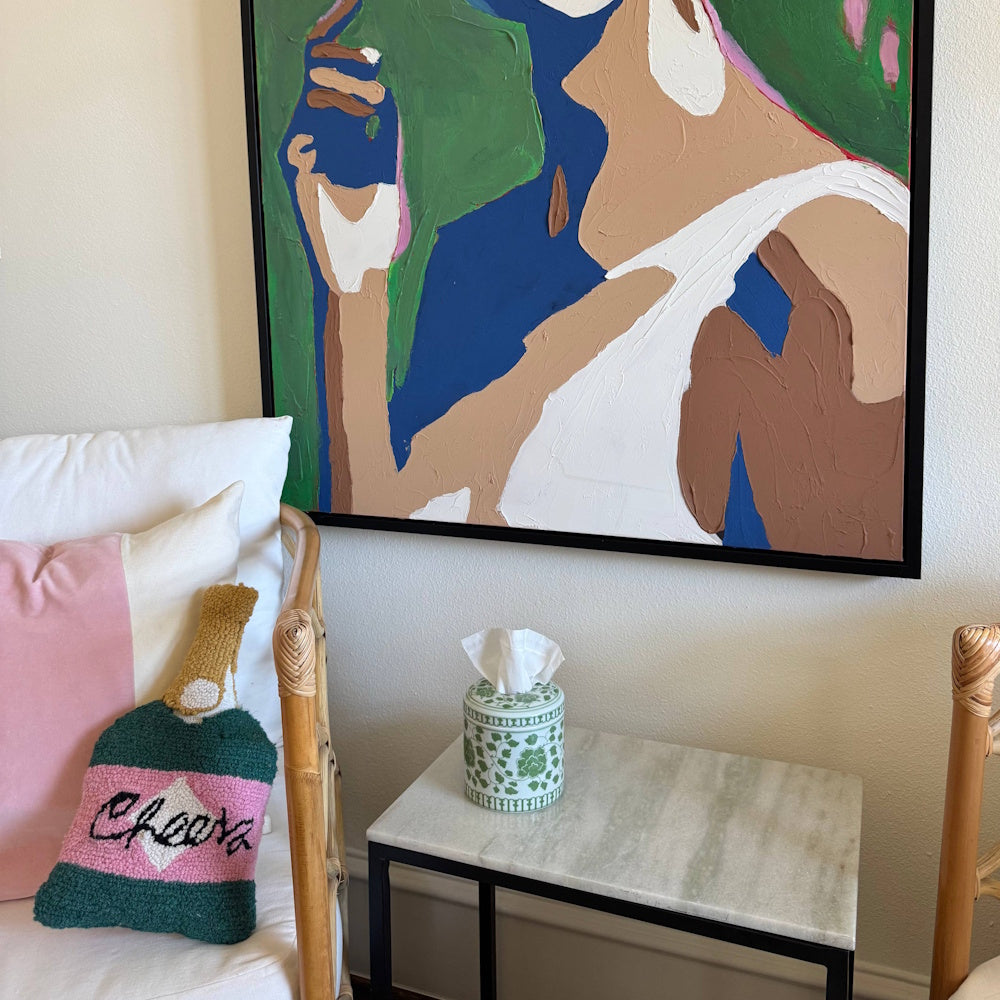 Green and white floral tissue vessel on a marble-topped side table below a colorful abstract painting.