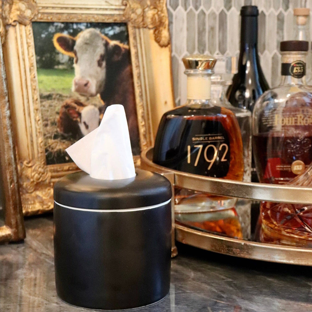 Matte black tissue dispenser in front of framed cow art and liquor bottles on a bar counter.
