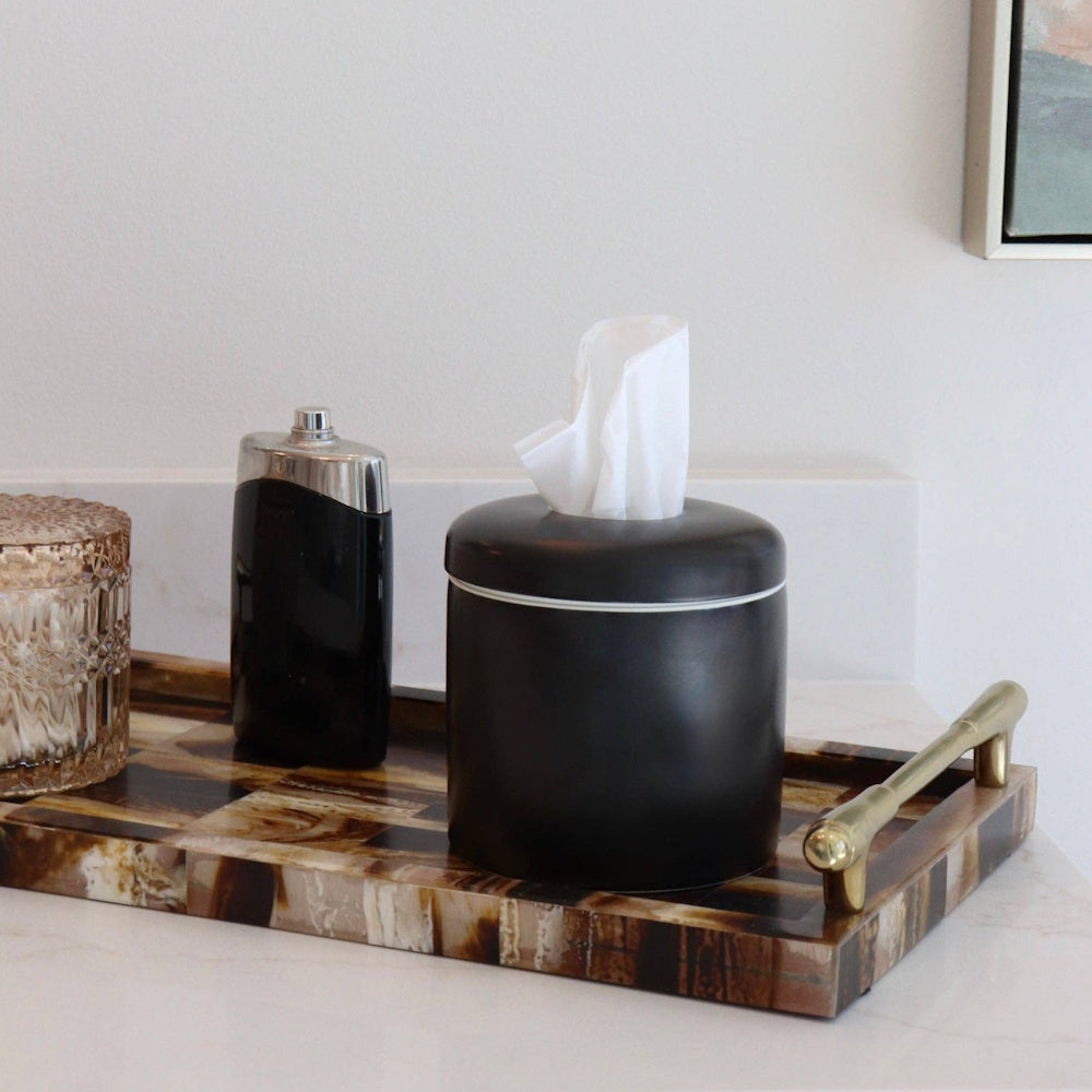 Matte black tissue dispenser on a brown patterned tray next to a perfume bottle.