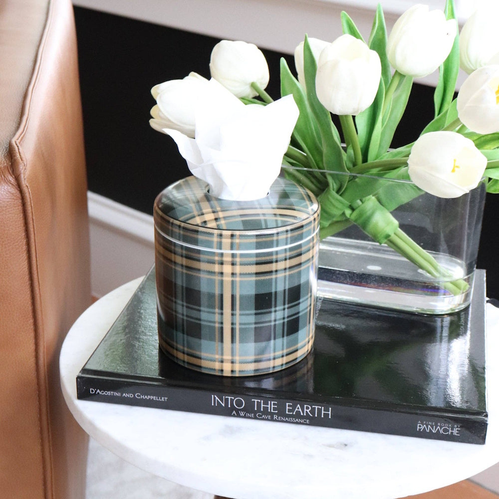 A plaid tissue box cover on a black book next to white tulips in a glass vase on a marble side table.