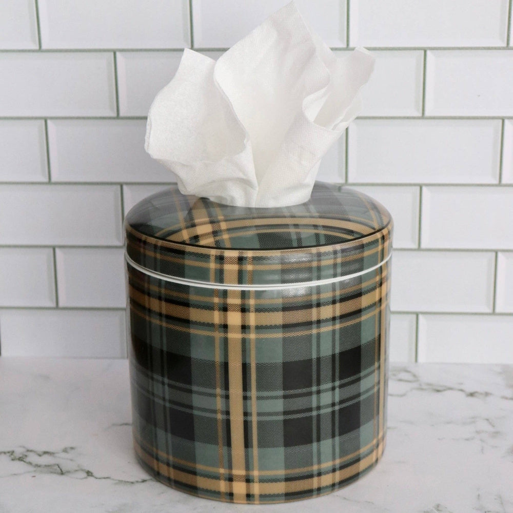 A cylindrical plaid tissue box cover with a white tissue on a marble counter against a white subway tile wall.