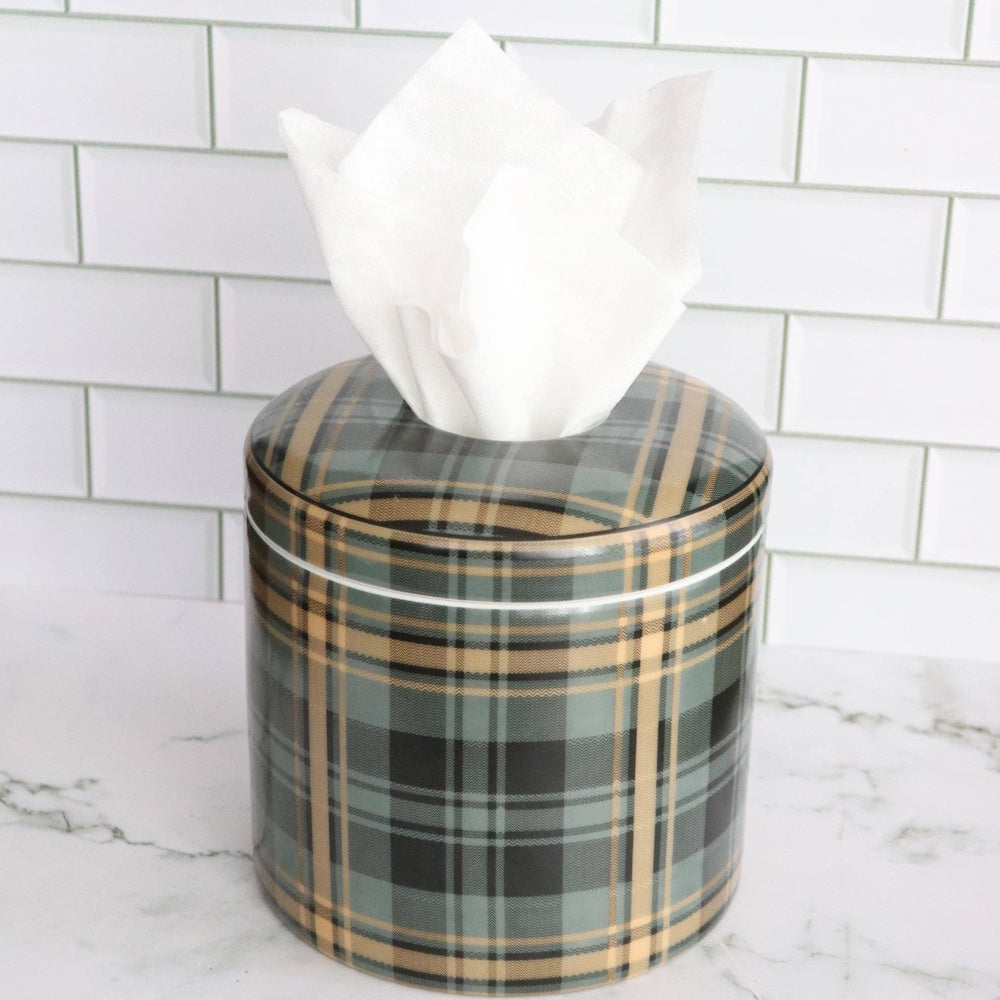 A cylindrical green and tan plaid tissue box cover on a marble surface with a white subway tile background.