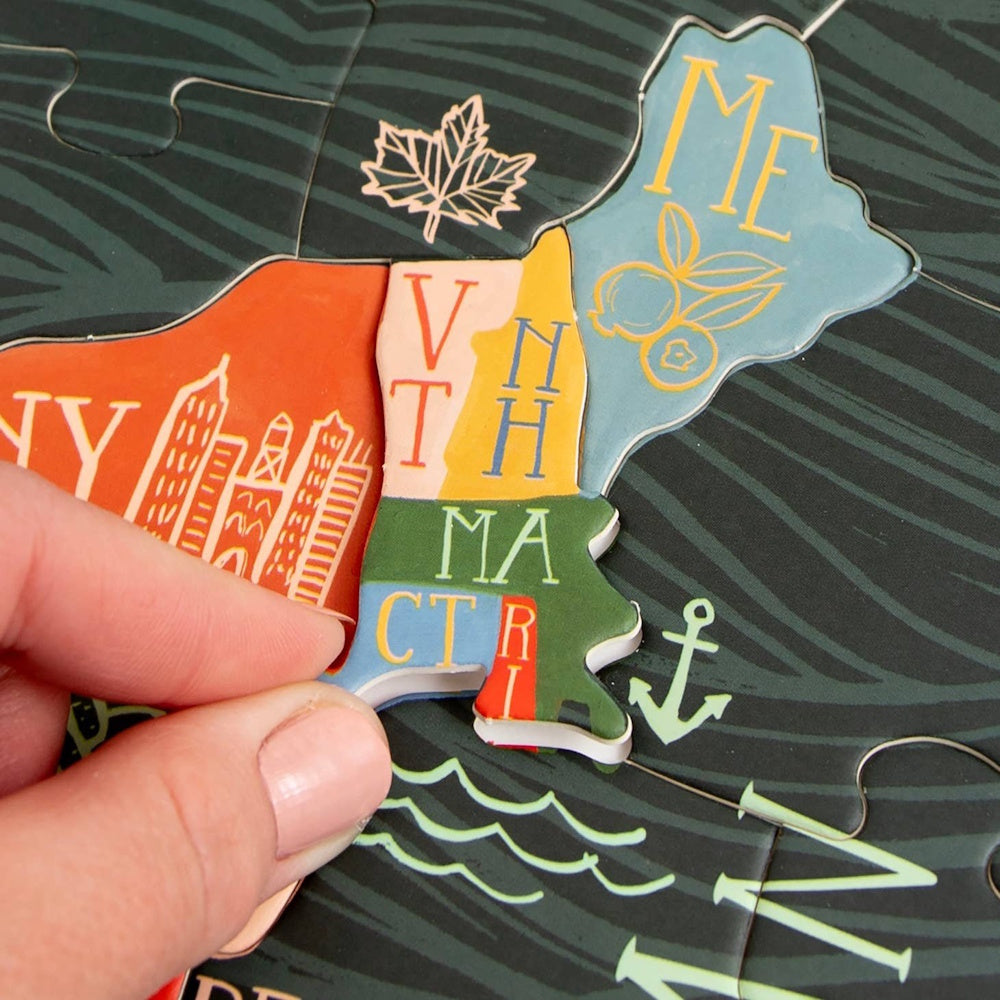 A person's hand places a colorful New England puzzle piece into a United States map puzzle with stylized icons.