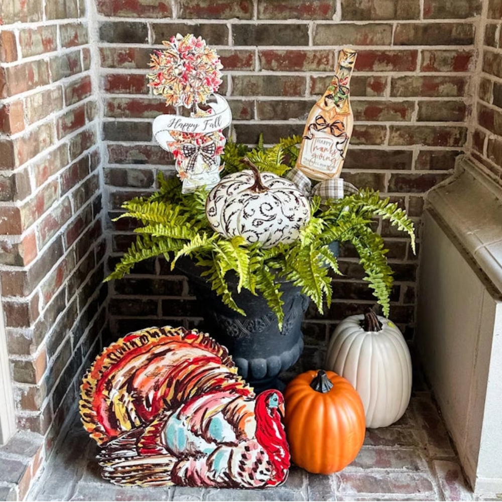 Fall porch display with a colorful turkey sign, pumpkins, and autumn decorations in front of a brick wall.