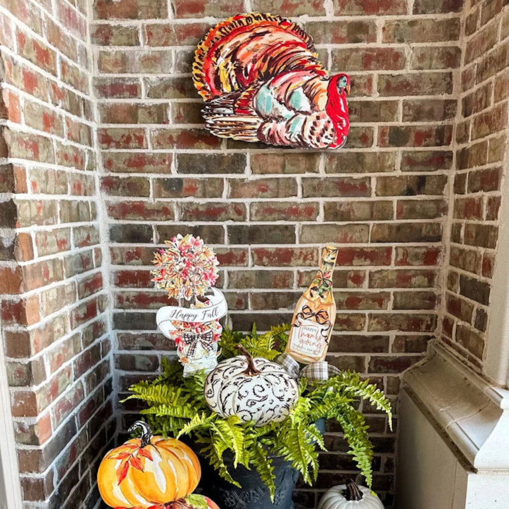 Thanksgiving porch decorations with a colorful turkey sign on a brick wall above pumpkins and ferns.