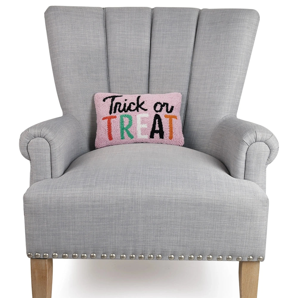 Pink "Trick or Treat" hooked throw pillow displayed on a gray tufted armchair with nailhead trim.