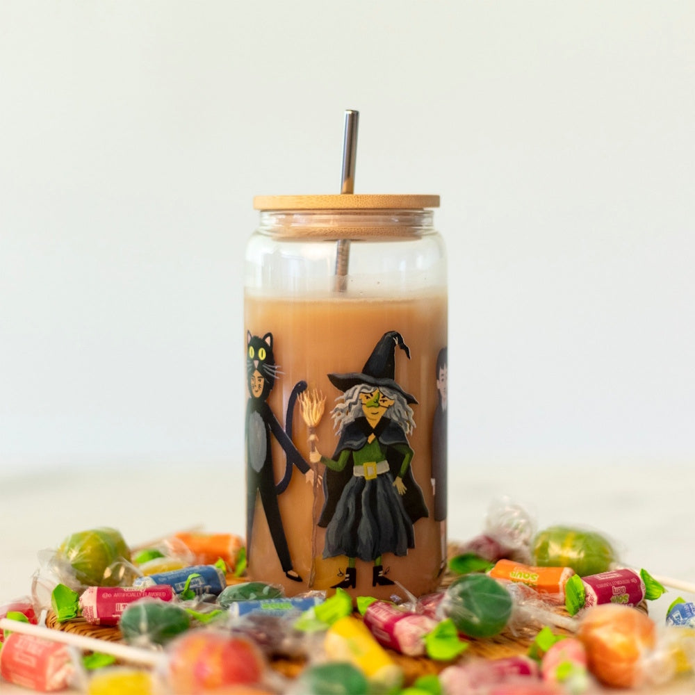 A Halloween-themed glass can cup filled with iced coffee, surrounded by scattered candy pieces.