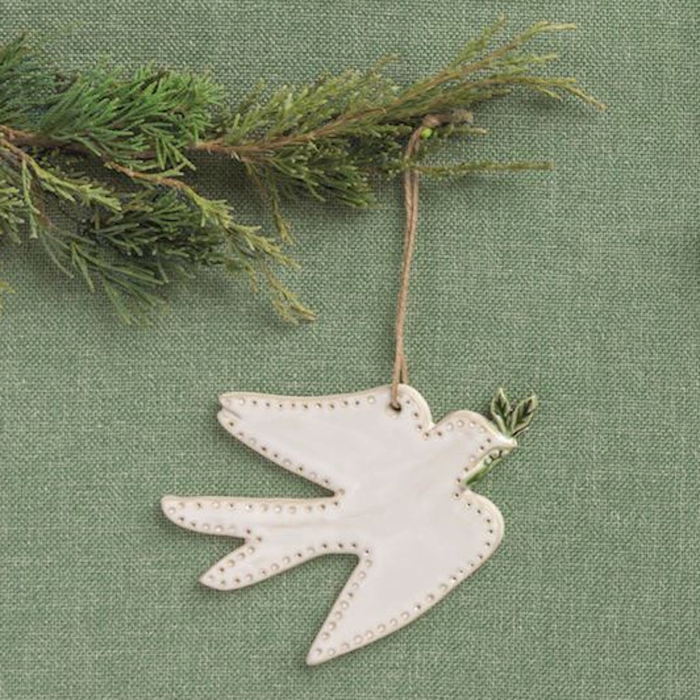 White ceramic dove ornament holding a green sprig, hanging by a twine string against a green background.