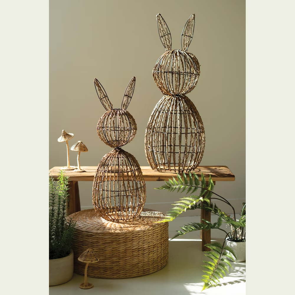 Two woven twig and wire rabbit sculptures on a wooden bench with wicker mushrooms and potted ferns in a bright room.